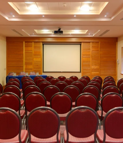 Empty hotel conference room with rows of red chairs facing a projector screen, warm wood walls, ready for a family event or meeting