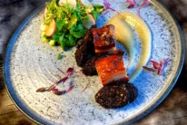 Family enjoying a plated gourmet pork belly dish with puree and fresh salad in a cosy restaurant setting (78 characters)