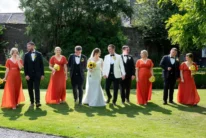 Bride and groom walking with wedding party on sunny hotel garden lawn, bridesmaids in red with sunflowers