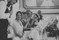 Bride smiling at groom giving a speech while family applaud at a hotel wedding breakfast