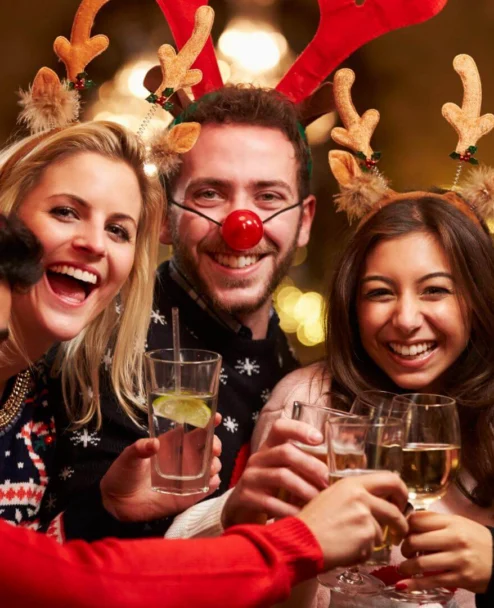 Family group in festive jumpers and antler headbands cheers with drinks in a warmly lit hotel lounge