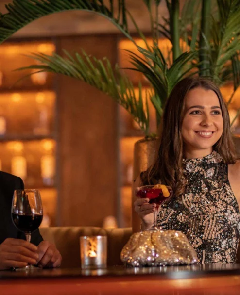 Smiling couple enjoying cocktails in a warm hotel bar lounge with soft candlelight and plants